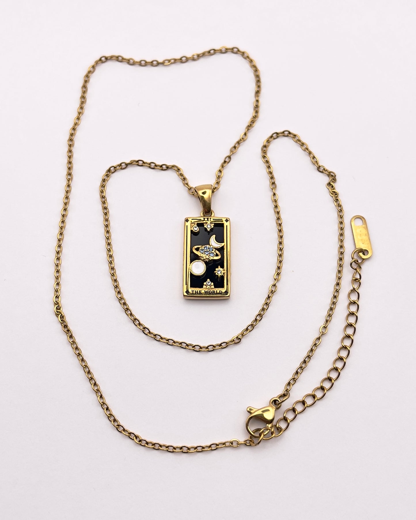 Full front view of an enchanting gold plated necklace perfected with a rectangular tarot card, the World (-XXI-), sculpted with stunning gold plated artistry, detailed with white and blue zircons, white enamel, and black stone backed.