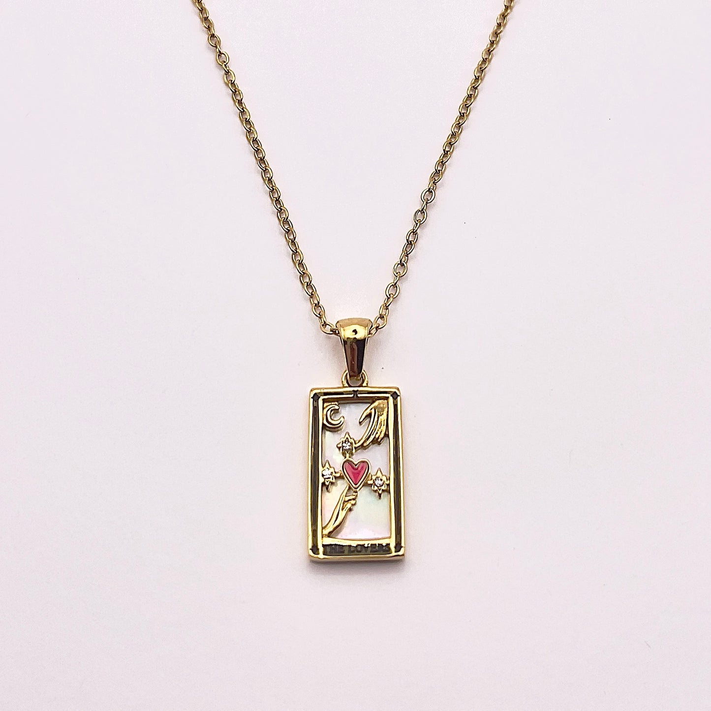 Front view of an enchanting gold plated necklace perfected with a rectangular tarot card, the Lovers (-X-), sculpted with stunning gold plated artistry, detailed with white zircons and red enamel, and white pearl colored stone backed.