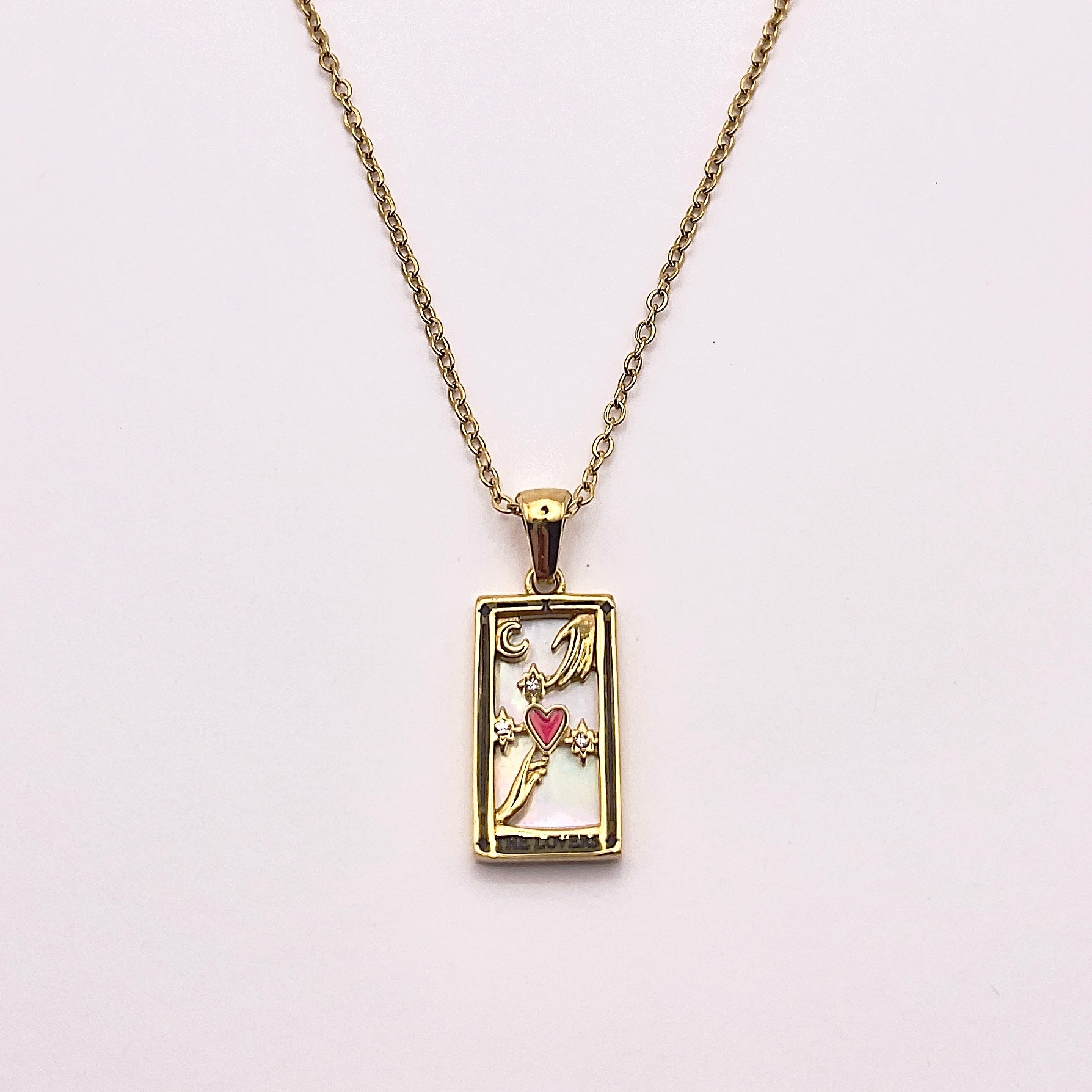 Front view of an enchanting gold plated necklace perfected with a rectangular tarot card, the Lovers (-X-), sculpted with stunning gold plated artistry, detailed with white zircons and red enamel, and white pearl colored stone backed.