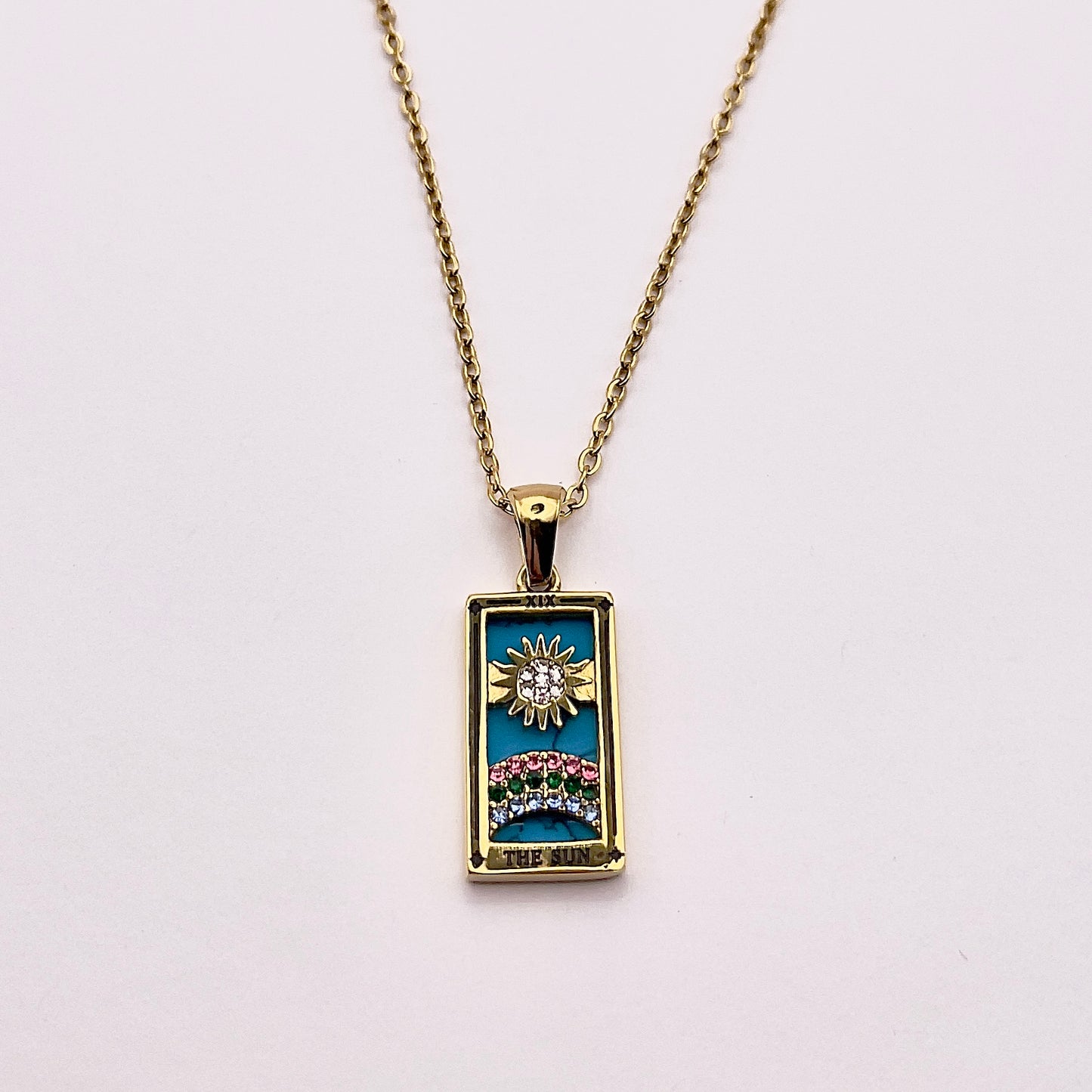 Front view of an enchanting gold plated necklace perfected with a rectangular tarot card, the Sun (-XIX-), sculpted with stunning gold plated artistry, detailed with pink, green, and white zircons, and blue lapis lazuli replica stone backed.