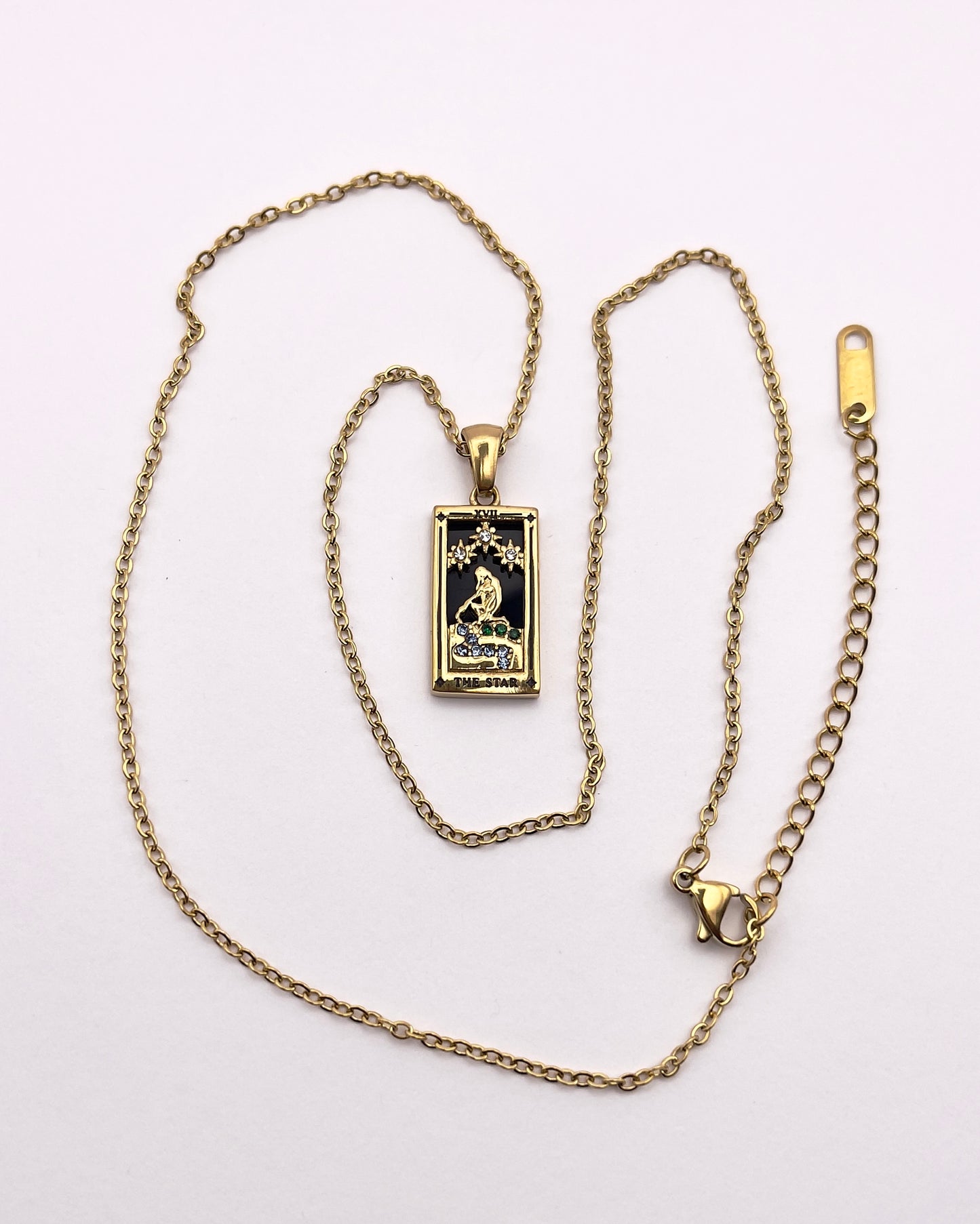 Full front view of an enchanting gold plated necklace perfected with a rectangular tarot card, the Star (-XVII-), sculpted with stunning gold plated artistry, detailed with white and green zircons, and black stone backed.