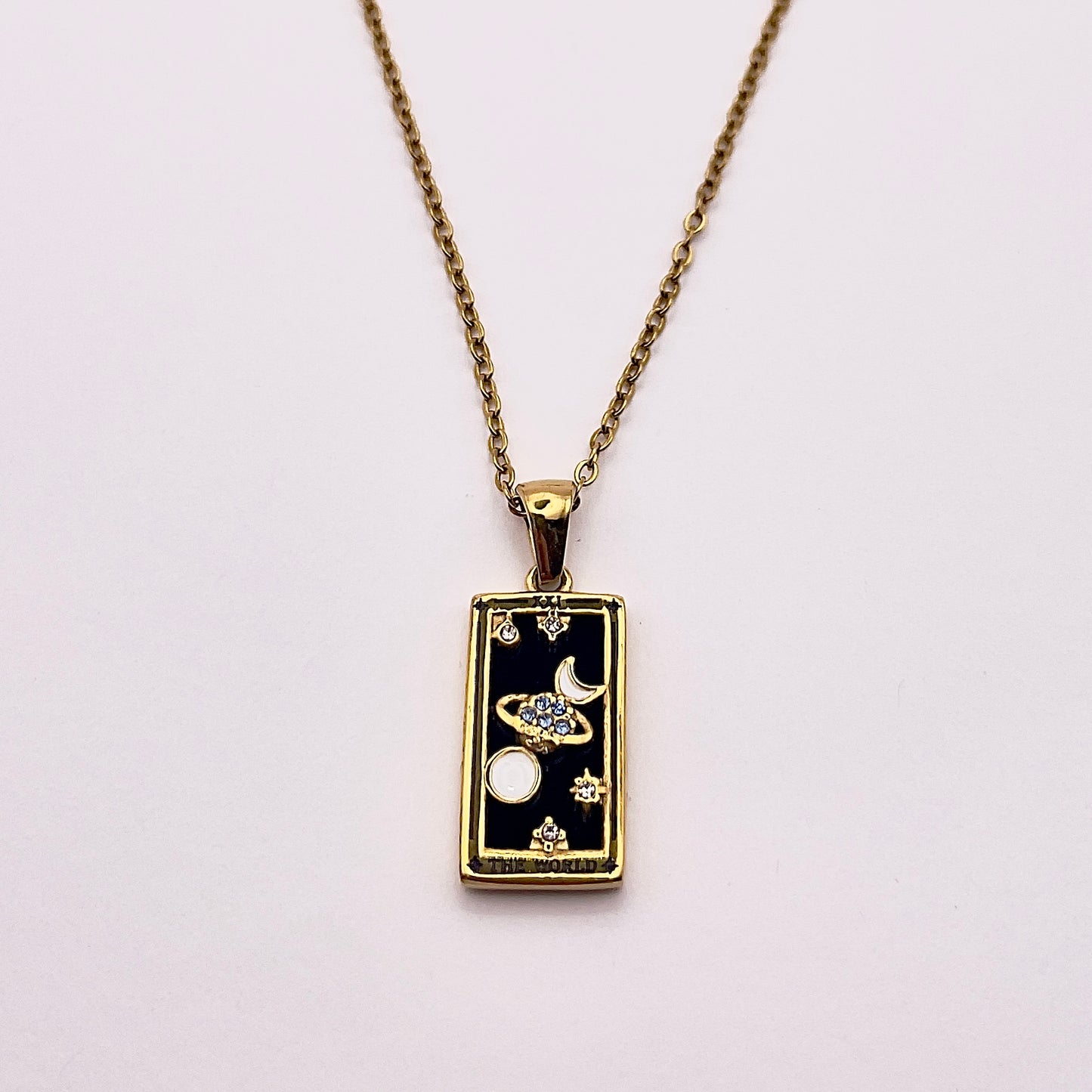 Front view of an enchanting gold plated necklace perfected with a rectangular tarot card, the World (-XXI-), sculpted with stunning gold plated artistry, detailed with white and blue zircons, white enamel, and black stone backed.