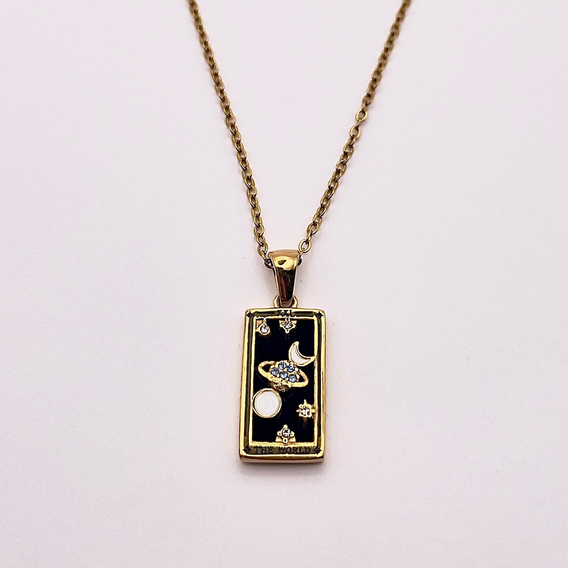 Front view of an enchanting gold plated necklace perfected with a rectangular tarot card, the World (-XXI-), sculpted with stunning gold plated artistry, detailed with white and blue zircons, white enamel, and black stone backed.