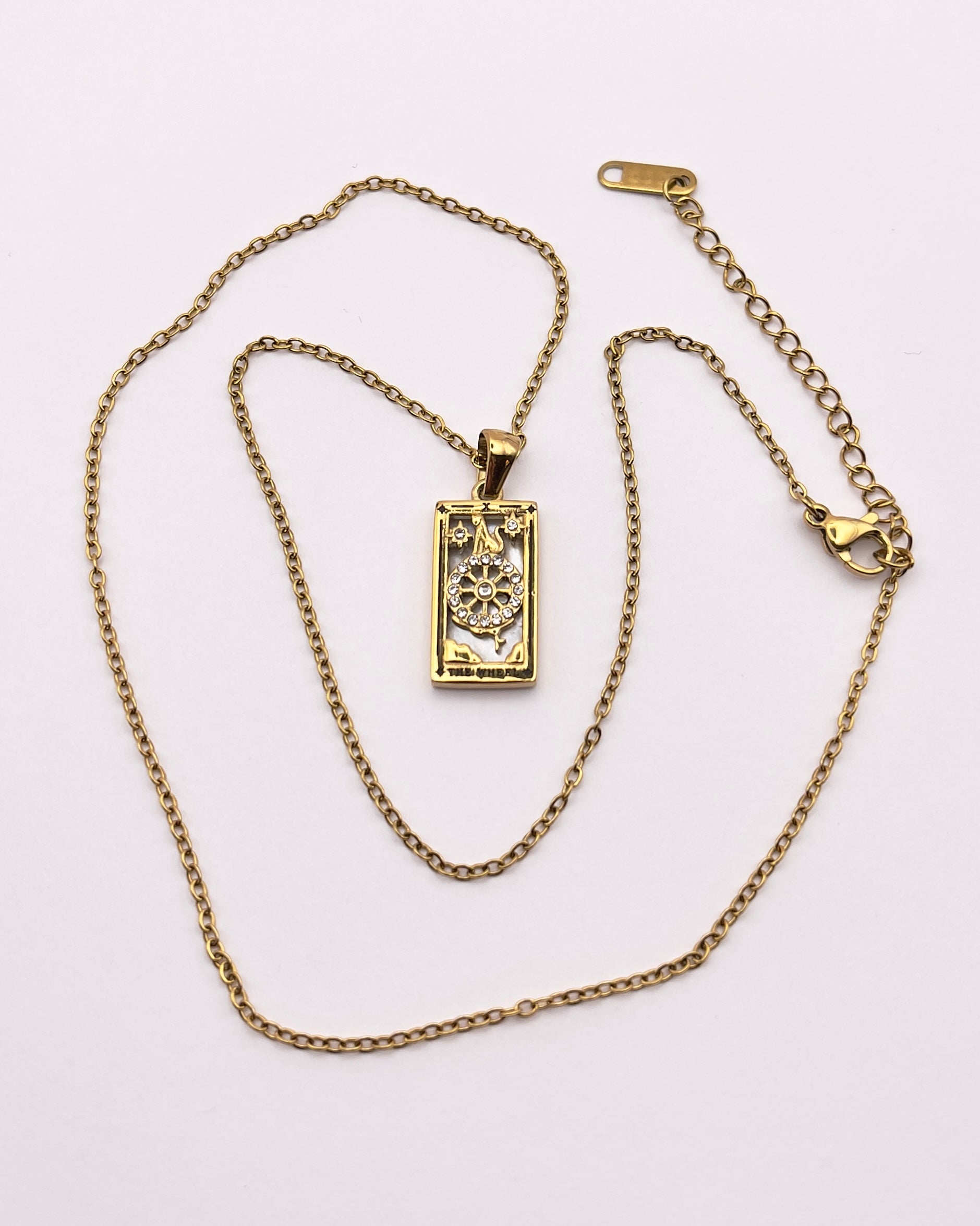 Full front view of an enchanting gold plated necklace perfected with a rectangular tarot card, the Wheel (-X-), sculpted with stunning gold plated artistry, detailed with white zircons, and white pearl colored stone backed.