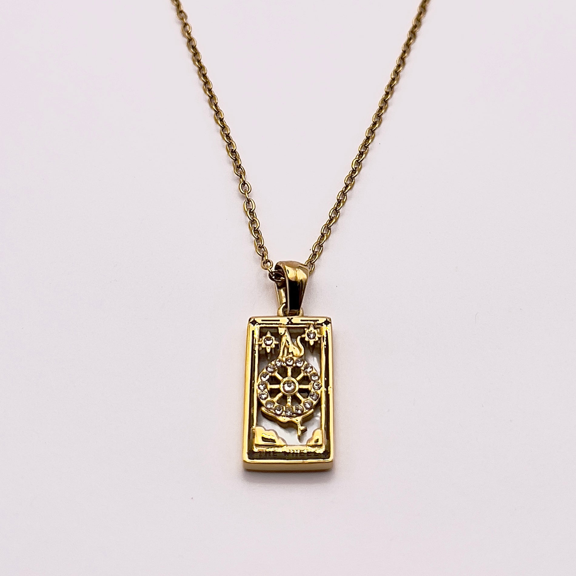 Front view of an enchanting gold plated necklace perfected with a rectangular tarot card, the Wheel (-X-), sculpted with stunning gold plated artistry, detailed with white zircons, and white pearl colored stone backed.