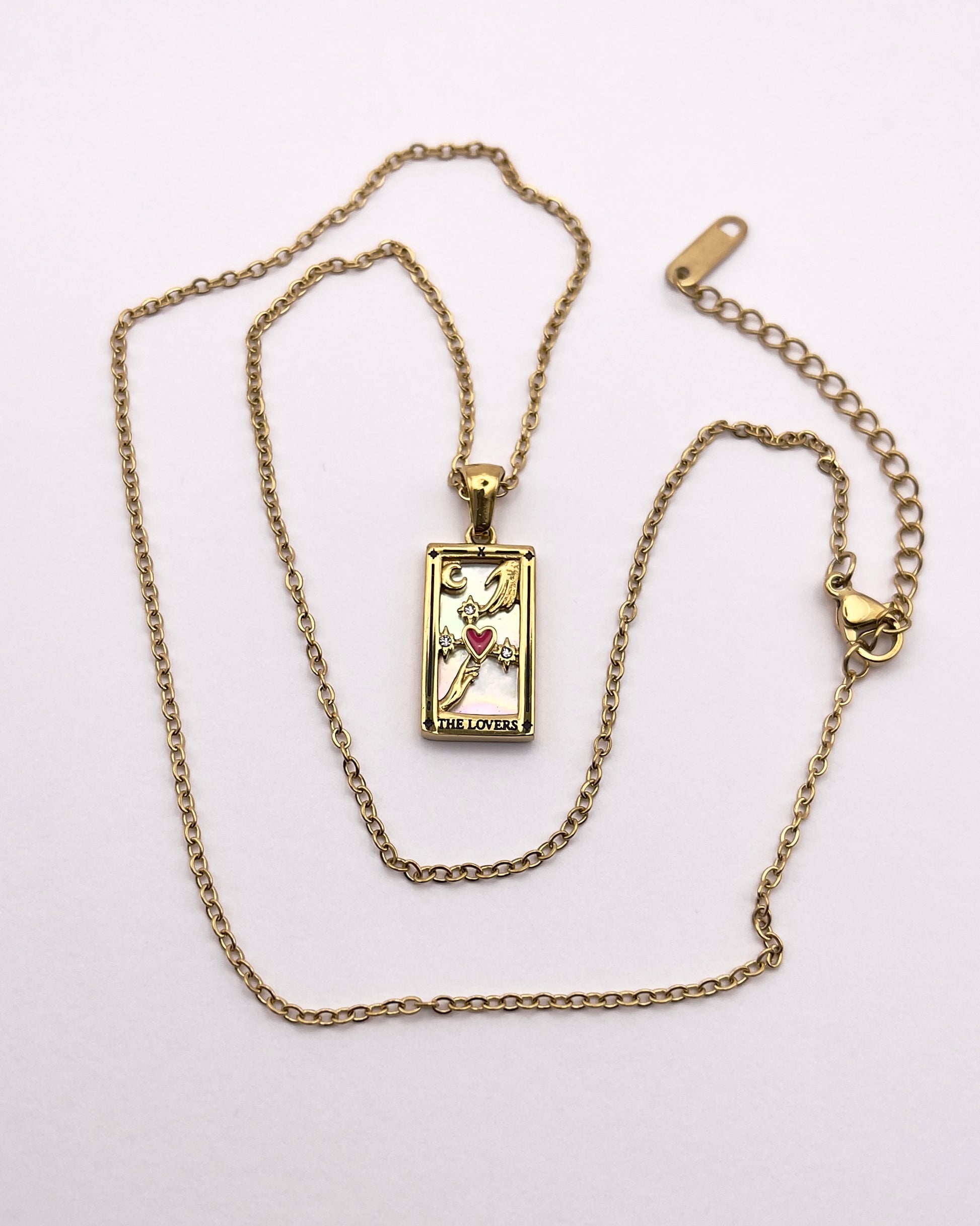 Full front view of an enchanting gold plated necklace perfected with a rectangular tarot card, the Lovers (-X-), sculpted with stunning gold plated artistry, detailed with white zircons and red enamel, and white pearl colored stone backed.