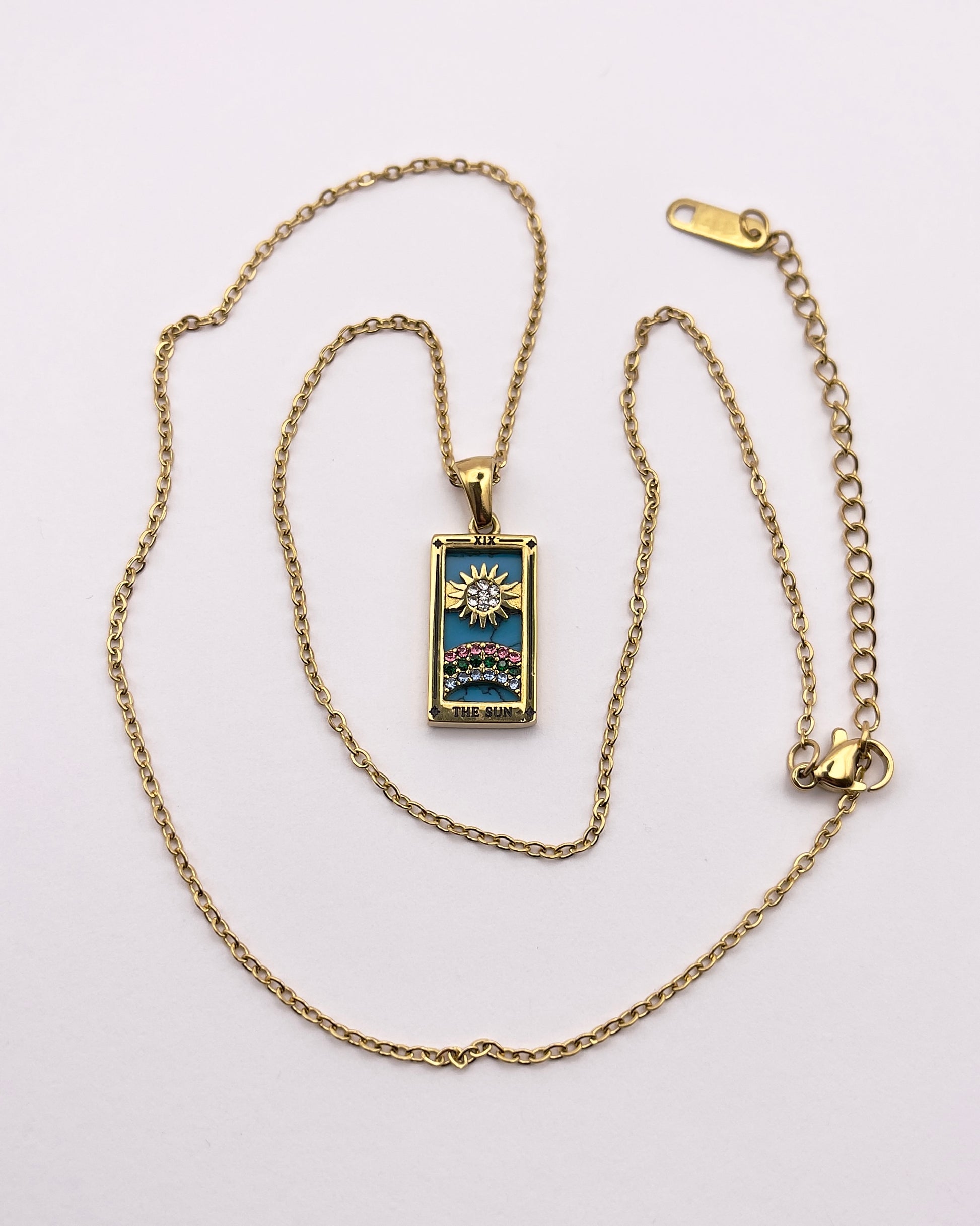 Full front view of an enchanting gold plated necklace perfected with a rectangular tarot card, the Sun (-XIX-), sculpted with stunning gold plated artistry, detailed with pink, green, and white zircons, and blue lapis lazuli replica stone backed.