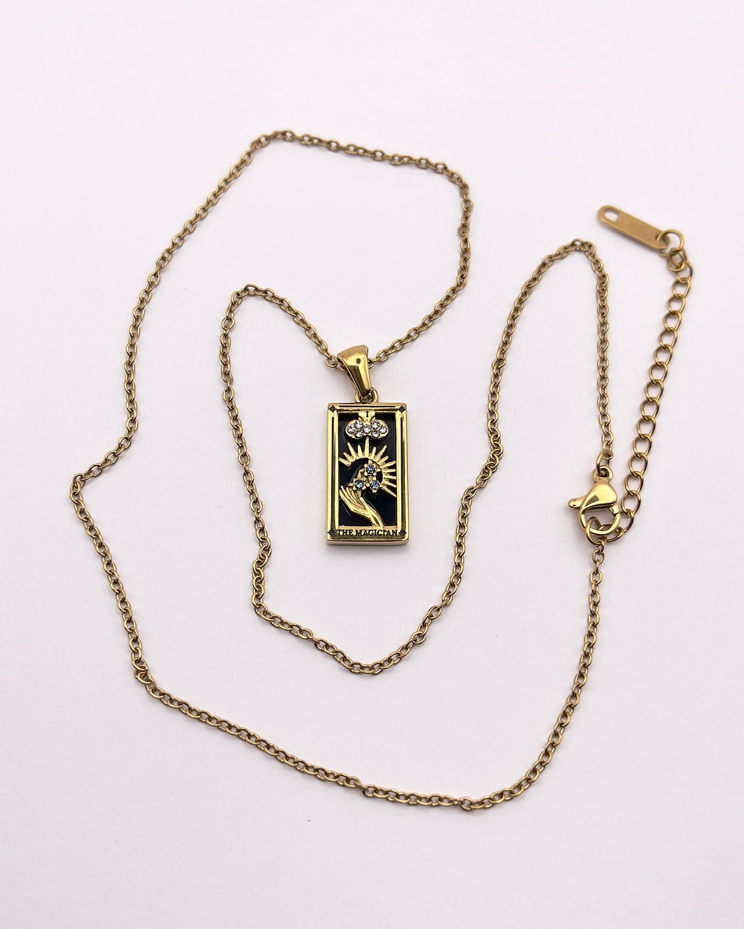 Full front view of an enchanting gold plated necklace perfected with a rectangular tarot card, the Magician (-I-), sculpted with stunning gold plated artistry, detailed with white and blue zircons, and black stone backed.